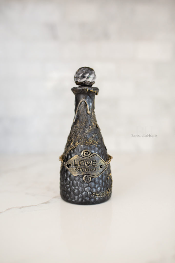 Close up view of the Love potion bottle. It has drips made out of mod podge and baking soda and lots of texture that is dry brushed gold and silver. The potion bottle has a polymer clay tag that’s attached to a chain that reads Love potion.