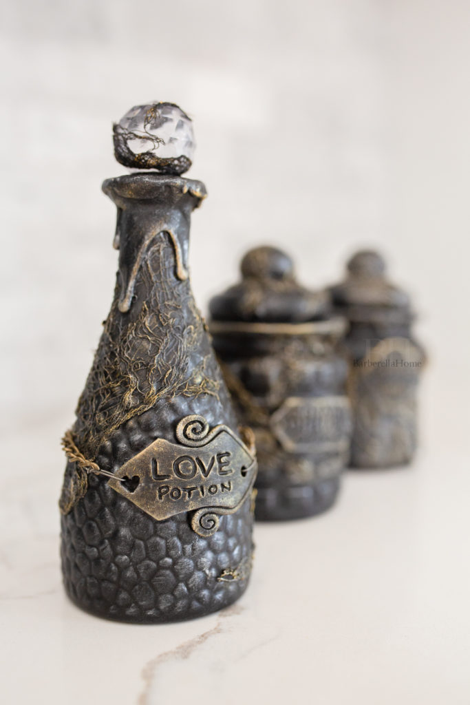 Close up view of the Love potion bottle. It has drips made out of mod podge and baking soda and lots of texture that is dry brushed gold and silver. The potion bottle has a polymer clay tag that’s attached to a chain that reads Love potion. 