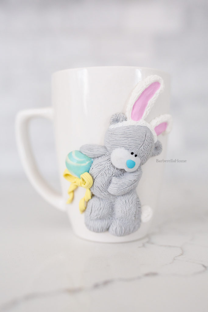 Mug art: 3D grey tatty teddy bear dressed as a bunny for easter. Holding a blue eggs with yellow ribbon. Made out of polymer clay.