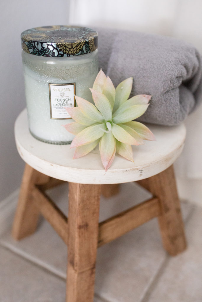 Little foot stool from Hobby that has white top and wooden legs with candle, faux succulent, and towel for decor.