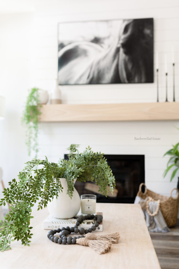 Pull back view of decor on coffee table with fireplace in the background. White ceramic pot (from walmart) with greenery (from hobby lobby) hanging over a coffee book (Home body by Joanna Gaines), candle (tides candle), and grey wood beads (from Hobby lobby). 