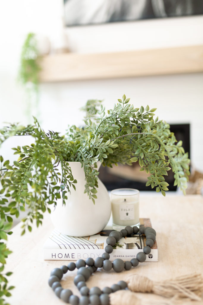 Closeup of decor on coffee table. White ceramic pot (from walmart) with greenery (from hobby lobby) hanging over a coffee book (Home body by Joanna Gaines), candle (tides candle), and grey wood beads (from Hobby lobby). 