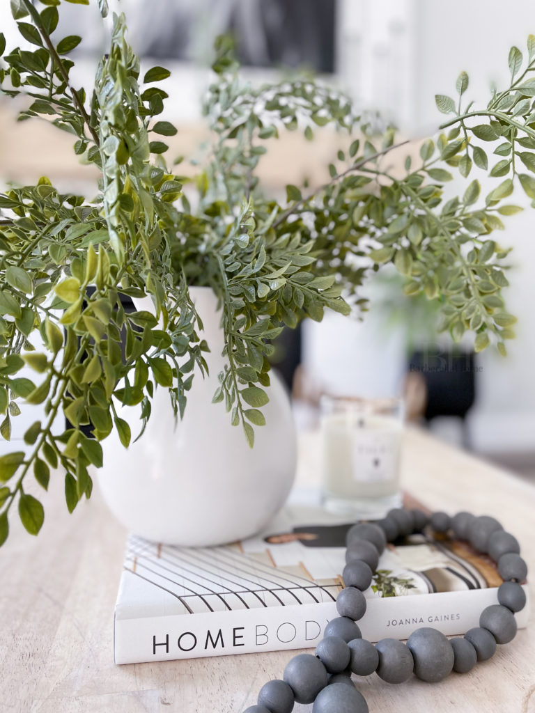 Closeup of decor on coffee table. White ceramic pot (from walmart) with greenery (from hobby lobby) hanging over a coffee book (Home body by Joanna Gaines), candle (tides candle), and grey wood beads (from Hobby lobby). 