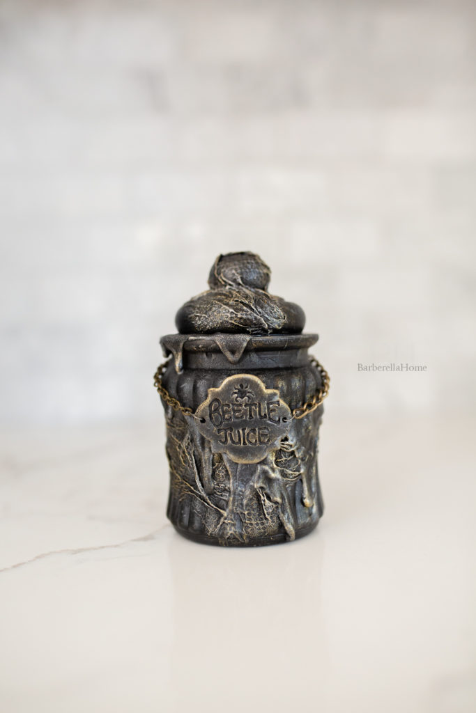 Close up view of the Beetle Juice potion bottle. It has thick drips made out of mod podge and baking soda and lots of texture that is dry brushed gold and silver. The potion bottle has a polymer clay tag that’s attached to a chain that reads Beetle Juice.