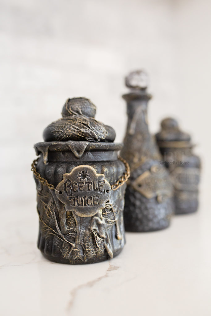 Close up view of the Beetle Juice potion bottle. It has thick drips made out of mod podge and baking soda and lots of texture that is dry brushed gold and silver. The potion bottle has a polymer clay tag that’s attached to a chain that reads Beetle Juice.