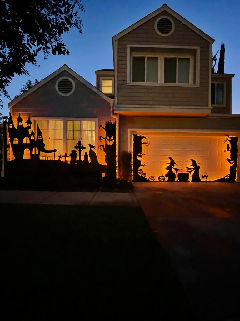 Step back view of the front of the house displaying the DIY Silhouette Decor for halloween - the witches stirring their cauldron, jack-o-lanterns with spooky trees, haunted house & graveyard silhouettes, back lit with orange and purple flickering lights.